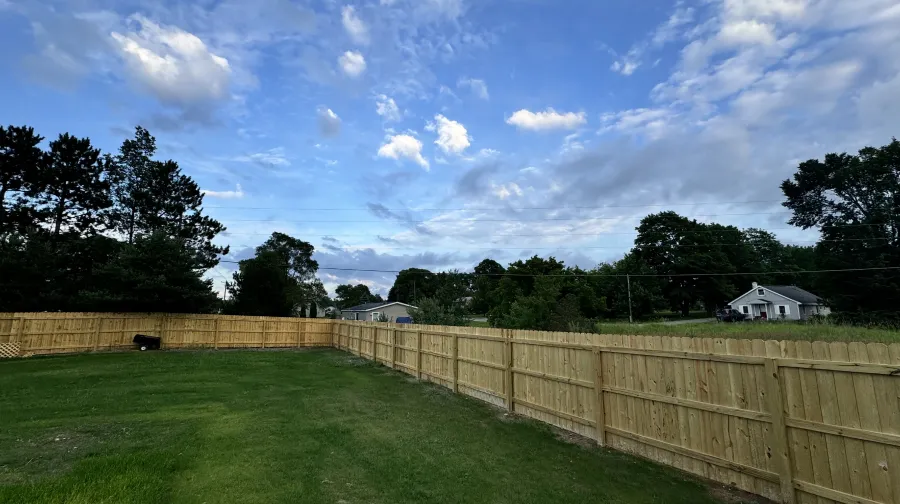 fence staining work le roy mi An empty backyard with a newly installed wooden privacy fence around the perimeter, under a partly cloudy sky.